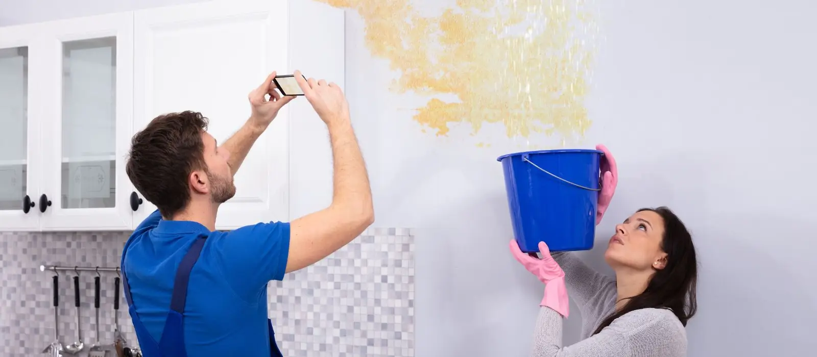 Man photographing a water-stained wall while a woman holds a bucket beneath the leak